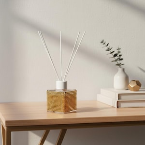 Könnte beinhalten: Ein dekorativer Reed-Diffusor mit einer quadratischen, goldfarbenen Basis, die mit kleinen Verzierungen bedeckt ist. Weiße Stäbchen ragen aus einer weißen, kreisförmigen Oberseite heraus. Der Diffusor steht auf einer hellbraunen Holzoberfläche.