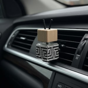 Könnte beinhalten: Ein Auto-Lufterfrischer mit geometrischem Design. Der untere Würfel ist mit schwarzen und silbernen Strasssteinen besetzt, während die Oberseite ein Holzquader mit zwei schwarzen Stäben ist. Der Lufterfrischer ist an einer Lüftungsöffnung im Auto befestigt.