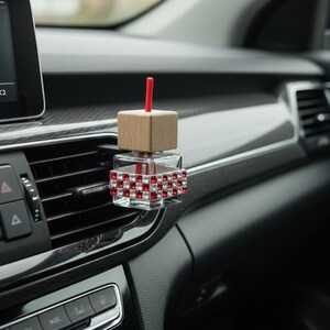 Könnte beinhalten: Ein Auto-Lufterfrischer mit einer klaren, quadratischen Glasflasche, verziert mit roten und weißen Kristallen. Er hat eine Holzoberseite und einen roten Stab. Der Lufterfrischer ist an einer Lüftungsöffnung im Auto befestigt und zeigt ein modernes Design.