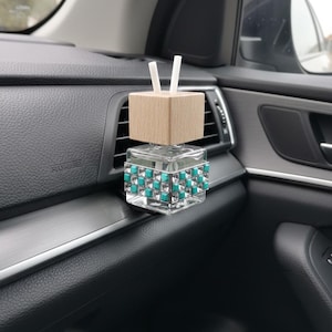 Könnte beinhalten: Ein Auto-Lufterfrischer mit einem transparenten Glasbehälter, verziert mit türkisfarbenen und transparenten quadratischen Akzenten. Er hat eine Holzoberseite mit zwei weißen Stäbchen. Der Lufterfrischer befindet sich auf einem Armaturenbrett.