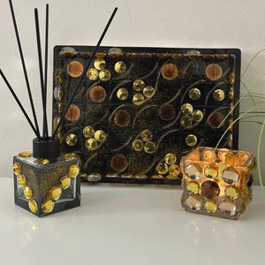 Könnte beinhalten: Ein dekoratives Set mit einem schwarzen Tablett, einem Glasdiffusor und einem Kerzenhalter. Das Tablett und der Kerzenhalter sind mit goldenen und braunen Verzierungen versehen. Der Diffusor hat schwarze Stäbchen und goldene Akzente.