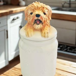 Op de afbeelding: Een witte keramische koekjespot met een bruin hondje op de deksel. De pot heeft pootafdrukontwerpen. De hond heeft een gedetailleerd gezicht met zwarte ogen en een roze tong. De pot staat op een houten oppervlak.