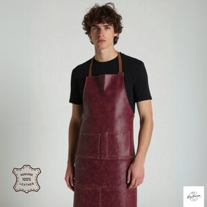 May include: A burgundy leather apron with brown leather straps and two large pockets. The apron has a distressed look and is made of genuine leather.