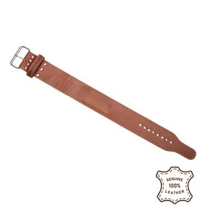 Peut inclure: Une ceinture d'halt&eacute;rophilie en cuir marron avec une boucle et des rivets argent&eacute;s. La ceinture a une forme rectangulaire avec une s&eacute;rie de trous pour le r&eacute;glage. Le coin inf&eacute;rieur droit de l'image porte un tampon indiquant "GENUINE 100% LEATHER."