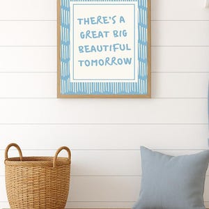 May include: A framed print with the words "THERE'S A GREAT BIG BEAUTIFUL TOMORROW" in blue lettering. The frame is wood-toned, and the background is white with blue accents. A woven basket and a blue pillow sit on a wooden bench below.