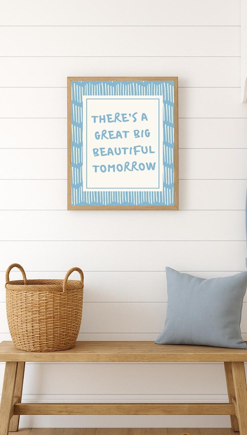 May include: A framed print with the words "THERE'S A GREAT BIG BEAUTIFUL TOMORROW" in blue lettering. The frame is wood-toned, and the background is white with blue accents. A woven basket and a blue pillow sit on a wooden bench below.