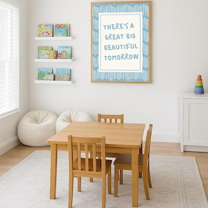 May include: A light wood children's table and chairs set in a bright room. A framed print with the text "THERE'S A GREAT BIG BEAUTIFUL TOMORROW" hangs on the wall. Bookshelves and bean bag chairs are also visible.