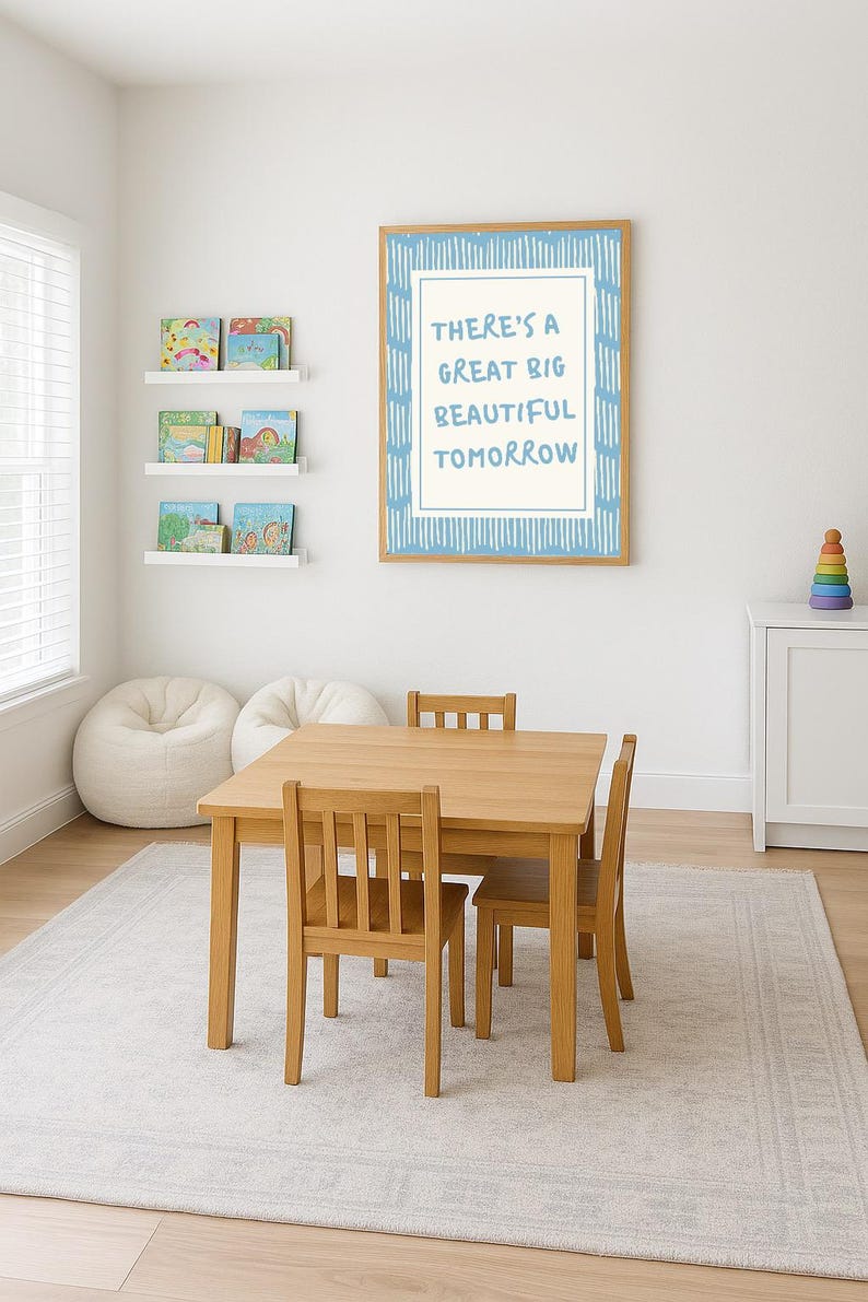 May include: A light wood children's table and chairs set in a bright room. A framed print with the text "THERE'S A GREAT BIG BEAUTIFUL TOMORROW" hangs on the wall. Bookshelves and bean bag chairs are also visible.