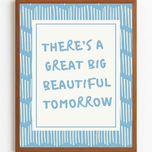 Op de afbeelding: Een ingelijste print met de tekst "THERE'S A GREAT BIG BEAUTIFUL TOMORROW" in lichtblauw op een witte achtergrond. De lijst is bruin en de print heeft een blauw-wit gestreepte rand.