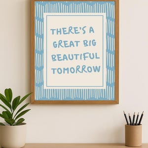 May include: Wooden framed art print with the text "THERE'S A GREAT BIG BEAUTIFUL TOMORROW" in blue. The print has a light blue and white design. The artwork is displayed on a wooden desk with a potted plant and pencils.