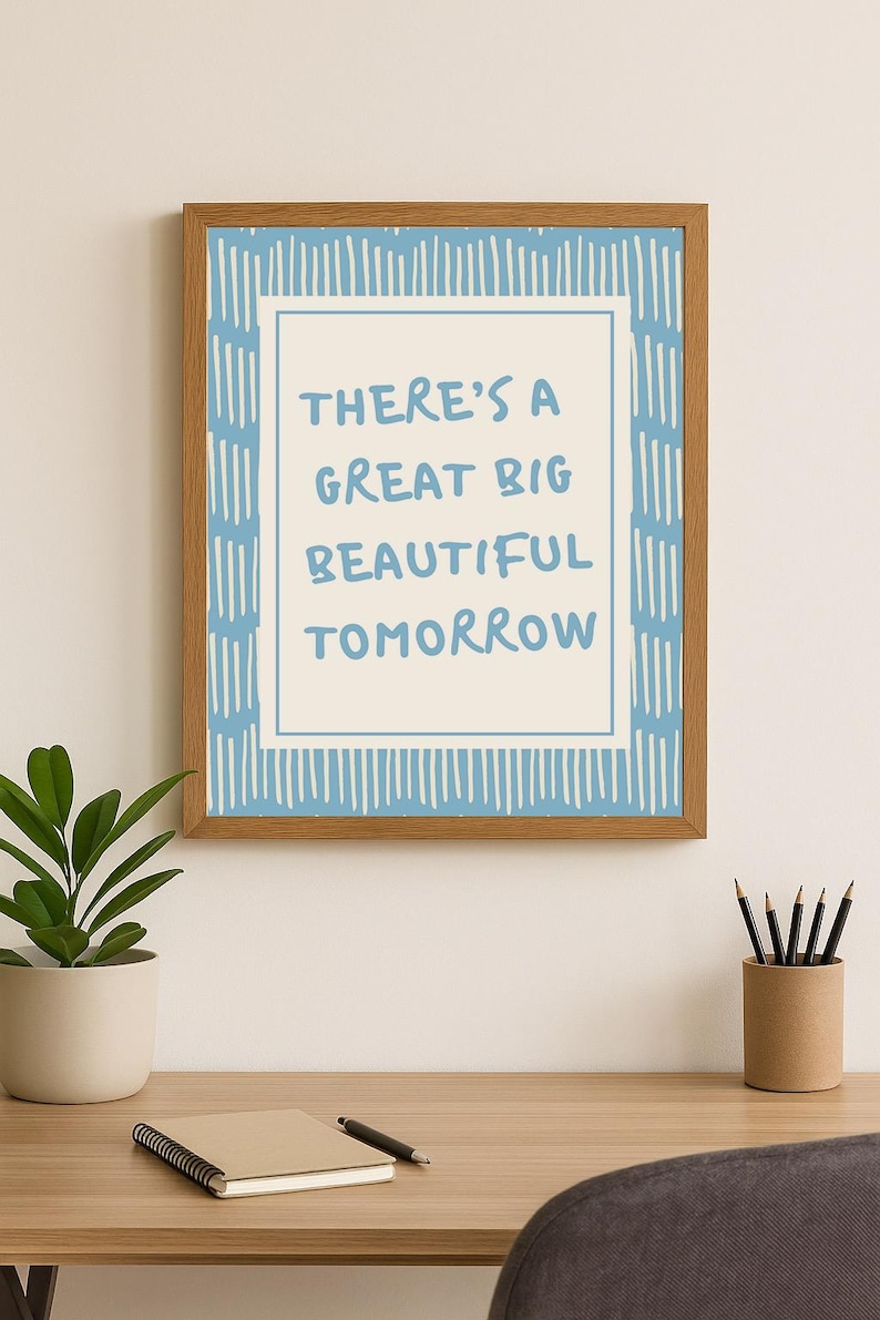 May include: Wooden framed art print with the text "THERE'S A GREAT BIG BEAUTIFUL TOMORROW" in blue. The print has a light blue and white design. The artwork is displayed on a wooden desk with a potted plant and pencils.