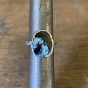May include: A silver ring with a colourful, irregularly shaped stone. The stone has blue, brown, and white hues, set in a silver bezel. The ring is displayed on a metal ring sizer, showcasing its design and craftsmanship.