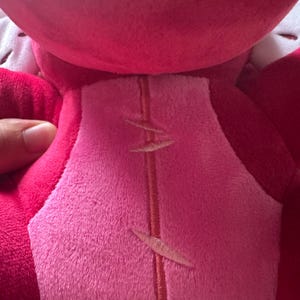 May include: Close-up of a plush toy in shades of pink and red. The toy's back features a lighter pink center panel with embroidered details. A hand is visible, holding the toy.