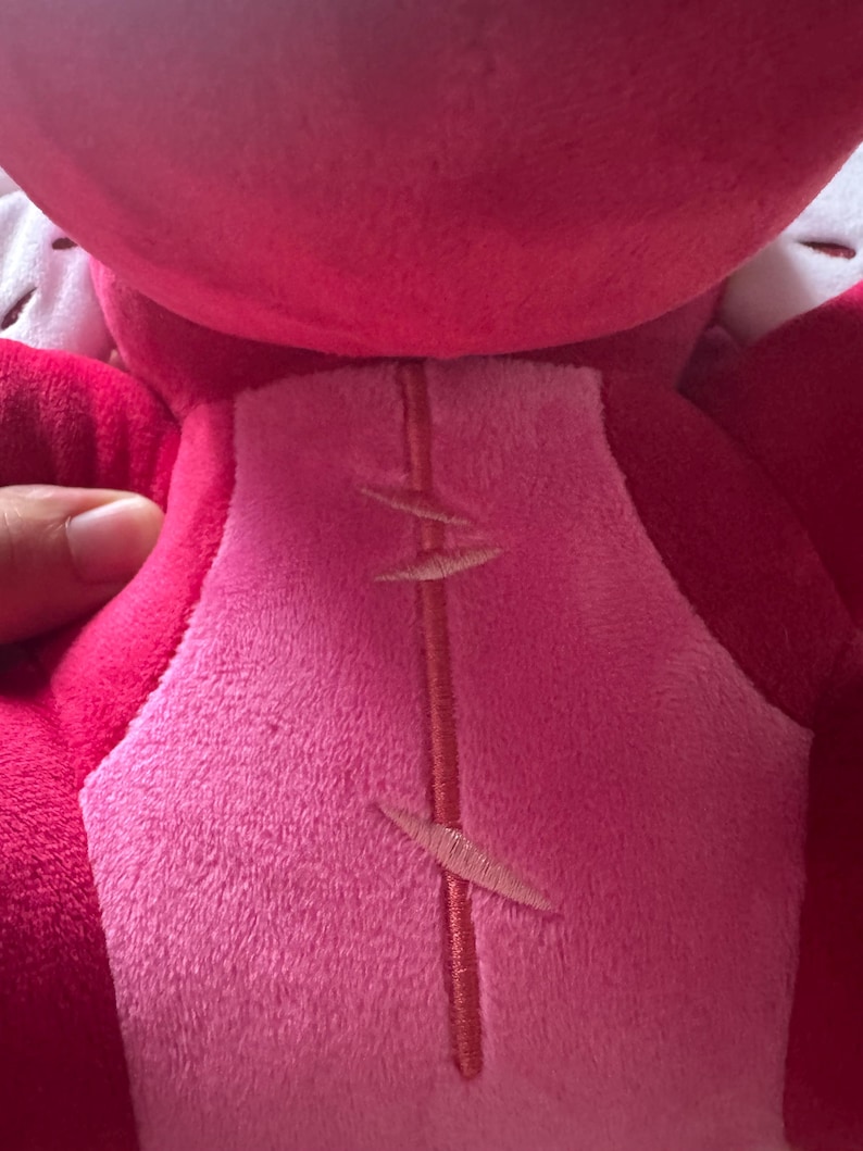 May include: Close-up of a plush toy in shades of pink and red. The toy's back features a lighter pink center panel with embroidered details. A hand is visible, holding the toy.