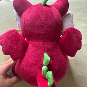 May include: A plush dragon toy in shades of red and green. The dragon has large ears, wings, and a tail with green accents. The toy is made of soft, plush material, and is designed for children.