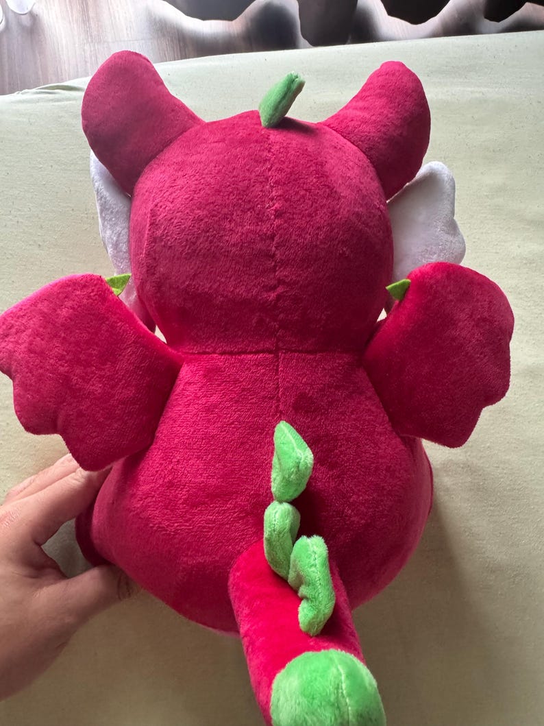 May include: A plush dragon toy in shades of red and green. The dragon has large ears, wings, and a tail with green accents. The toy is made of soft, plush material, and is designed for children.
