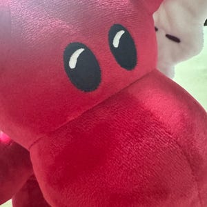 May include: Close-up of a plush red dragon toy with large black eyes and white highlights. The dragon's head and part of its body are visible, showcasing its soft, textured fabric. A white plush toy is partially visible in the background.
