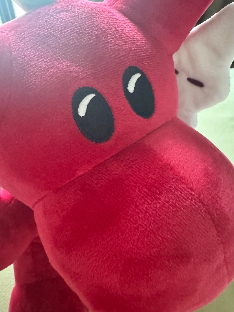 May include: Close-up of a plush red dragon toy with large black eyes and white highlights. The dragon's head and part of its body are visible, showcasing its soft, textured fabric. A white plush toy is partially visible in the background.