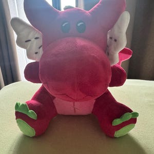 May include: A plush, red dragon toy with white wings and green accents on its feet. The dragon has large, round eyes and is sitting on a light green surface. The toy is made of soft material.