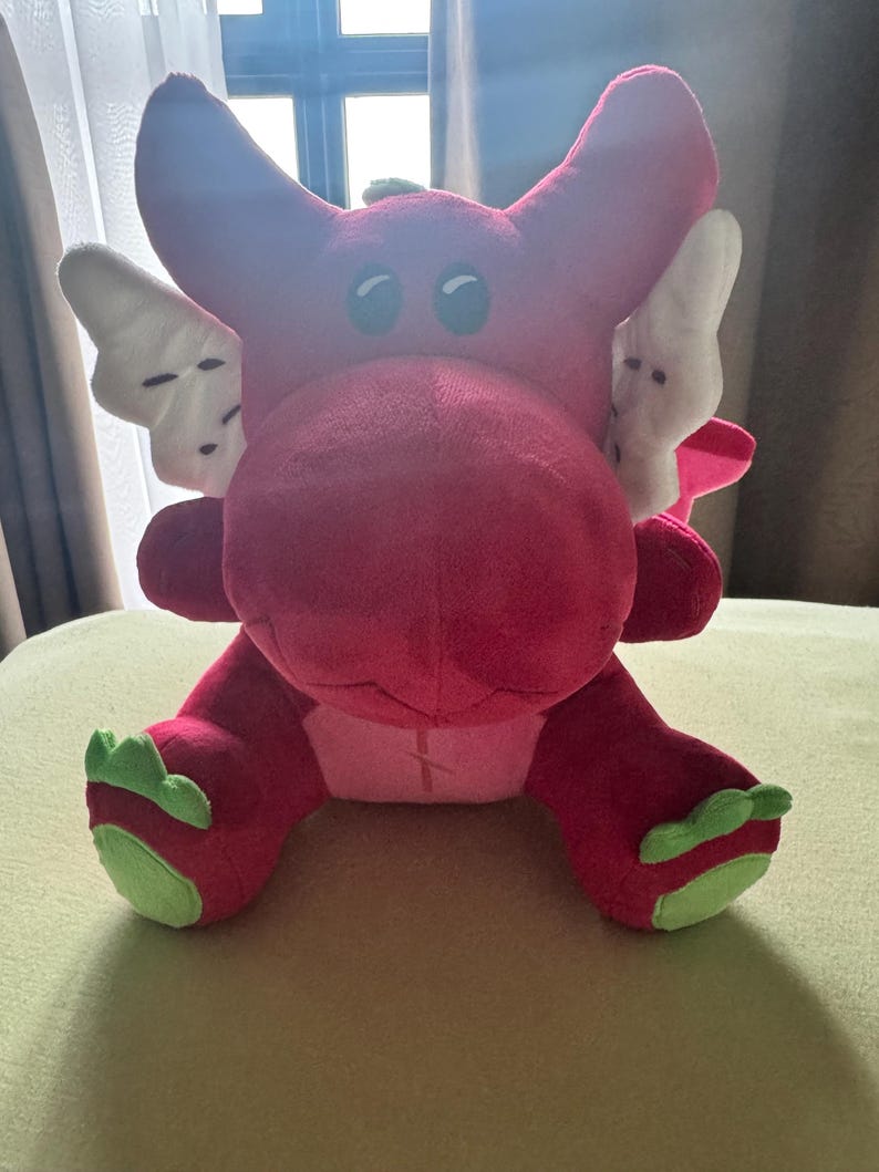 May include: A plush, red dragon toy with white wings and green accents on its feet. The dragon has large, round eyes and is sitting on a light green surface. The toy is made of soft material.