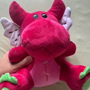 May include: A plush dragon toy in shades of pink and red, with white wings and green accents on the feet. The dragon has black eyes and a stitched design on its belly. A soft, cuddly children's toy.