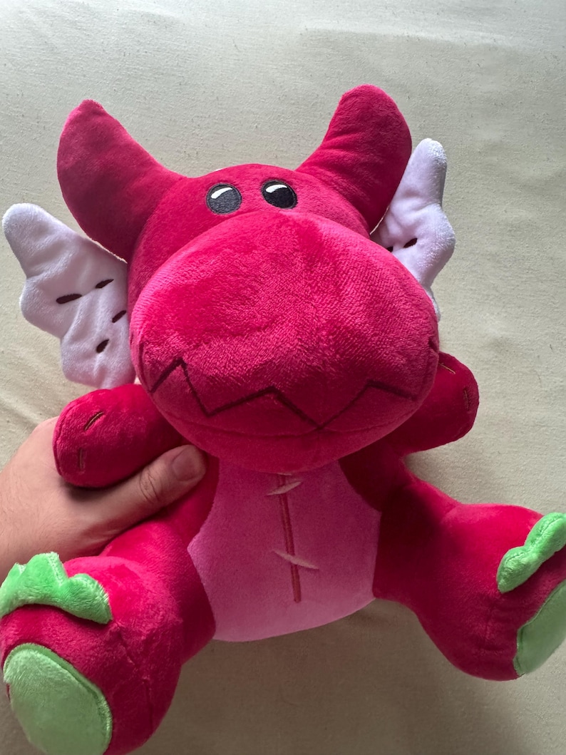 May include: A plush dragon toy in shades of pink and red, with white wings and green accents on the feet. The dragon has black eyes and a stitched design on its belly. A soft, cuddly children's toy.