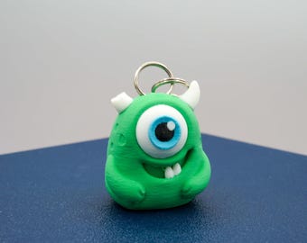 May include: A green monster keychain with one large eye, white horns, and a smiling mouth. The eye has a blue iris and black pupil. The keychain is made of a solid material and has a silver keyring.