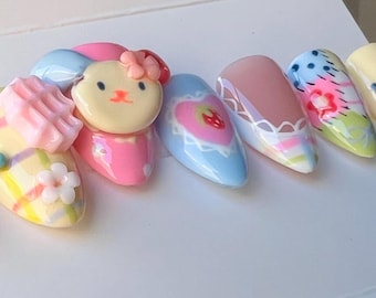 May include: A set of six pastel-colored artificial nails with various designs. The designs include a pink and white checkered pattern, a pink and blue heart with a strawberry, a light blue and white floral pattern, a pink and white floral pattern with polka dots, a light brown bunny with a pink bow, and a light brown and white floral pattern.