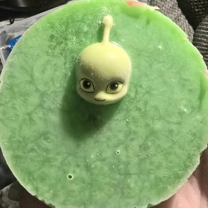 Puede incluir: Un jabón circular verde claro con una pequeña figura de alienígena amarillo pálido incrustada en el centro. El alienígena tiene ojos grandes y una pequeña sonrisa. El jabón tiene una superficie texturizada.