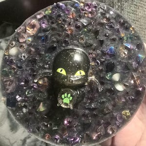 May include: A black cat figurine with yellow eyes and a green paw print, set in a dark, gem-studded resin base. The resin is filled with various colorful crystals and gems, creating a sparkling effect.