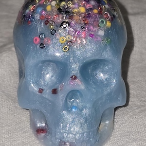 May include: A light blue skull-shaped decorative object with a glittery, bead-covered top. The skull has a smooth, glossy finish and is adorned with various colorful beads and glitter accents, creating a unique and eye-catching design.