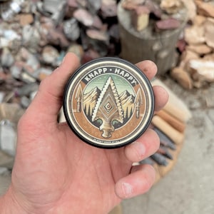May include: A round, black tin with the words "KNAPP HAPPY" encircling a central design. The design features a mountain range, an arrowhead, and the text "OR DON'T KNAPP AT ALL". The tin is held in a hand.