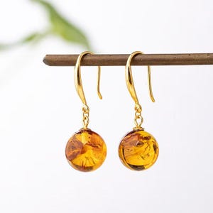 May include: A pair of gold-colored drop earrings with amber-colored, spherical pendants. The earrings are suspended from curved hooks and feature a warm, translucent glow. The amber orbs have natural inclusions, adding unique character to each earring.