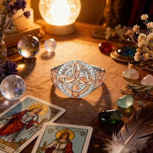May include: A silver ring with a Celtic knot design, accented with rose gold and clear gemstones. The ring is set against a backdrop of tarot cards, crystals, and candles, suggesting a mystical theme.