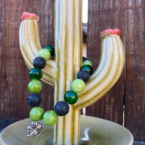 May include: A green and black beaded bracelet with a silver four-leaf clover charm displayed on a ceramic cactus stand.