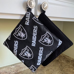 May include: Two black pot holders with a white Raiders logo and the word "RAIDERS" repeated on the fabric. The pot holders are hanging from hooks on a white cabinet.