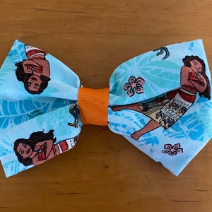 May include: A blue bow tie with a pattern of Moana and tropical flowers. The bow tie has an orange center.