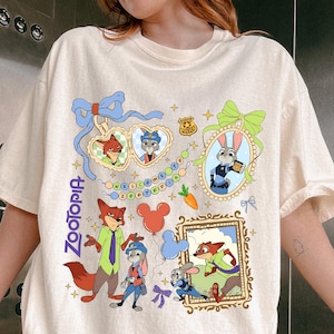 May include: Cream-colored t-shirt featuring a colorful Zootopia graphic. The design includes cartoon characters, frames, and the word "Zootopia" in blue. The shirt has short sleeves and a relaxed fit.
