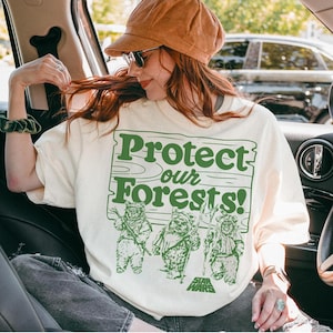 May include: A cream-coloured t-shirt with the green text "Protect our Forests!" and Ewok characters from Star Wars. The person is wearing a brown corduroy cap and sunglasses.