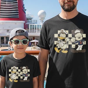 May include: Black t-shirts featuring a cartoon graphic of Donald Duck and Mickey Mouse in sailor and captain outfits. The characters are set against a checkered background. The shirts are worn by a child and an adult.