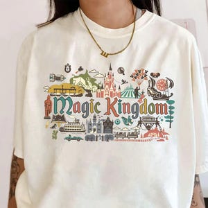 May include: Cream-colored t-shirt featuring a colorful graphic of the Magic Kingdom. The design includes various park icons and the words "Magic Kingdom" in a stylized font. The shirt has short sleeves and a relaxed fit.