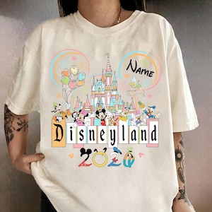 Op de afbeelding: Cr&egrave;mekleurig T-shirt met een kleurrijke Disneyland-afbeelding. Het ontwerp bevat een kasteel, ballonnen en diverse Disney-figuren. De tekst "Disneyland 2020" is prominent aanwezig.