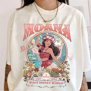 May include: Cream-colored t-shirt featuring a graphic of a woman in a floral crown, with the word "MOANA" in pink above her. The design includes flowers, butterflies, and the text "WALT DISNEY WORLD EST 2016".