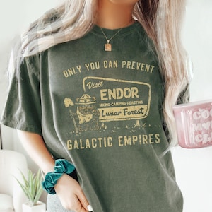 May include: A moss green t-shirt featuring the text "ONLY YOU CAN PREVENT Visit ENDOR Lunar Forest GALACTIC EMPIRES" in a retro design. The shirt is worn with grey shorts and a pink mug with the words "cool mom club".