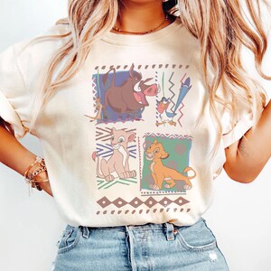 May include: Ivory-colored t-shirt featuring a colorful graphic of characters from The Lion King. The design includes images of Pumbaa, Timon, Simba, and Nala, set against geometric backgrounds. The word "IVORY" is printed at the bottom.