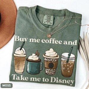 Puede incluir: Camiseta verde musgo con el texto "Buy me coffee and Take me to Disney". La camiseta presenta ilustraciones de cuatro bebidas de caf&eacute; diferentes. Un collar dorado est&aacute; drapeado sobre la camiseta.