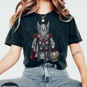 Op de afbeelding: Grafietkleurig T-shirt met een cartoonillustratie van Thor in een aan elkaar genaaid kostuum, met een pompoenemmer. Het ontwerp omvat een helm, cape en harnas. Het shirt is donkergrijs.