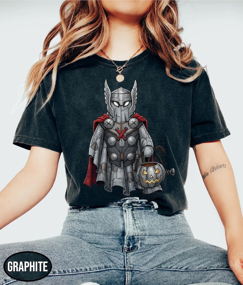 Op de afbeelding: Grafietkleurig T-shirt met een cartoonillustratie van Thor in een aan elkaar genaaid kostuum, met een pompoenemmer. Het ontwerp omvat een helm, cape en harnas. Het shirt is donkergrijs.