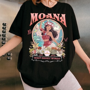 May include: Black t-shirt featuring the word "MOANA" in pink lettering above a circular graphic. The graphic depicts a woman in a floral crown and red outfit, surrounded by flowers and butterflies. The text "WALT DISNEY WORLD" is below.