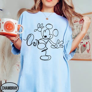 May include: Light blue t-shirt featuring a black line drawing of Mickey Mouse with the word "Cool" written in cursive. A white and orange mug with a sun design is held in the left hand. The word "Chambray" is printed on the bottom left.
